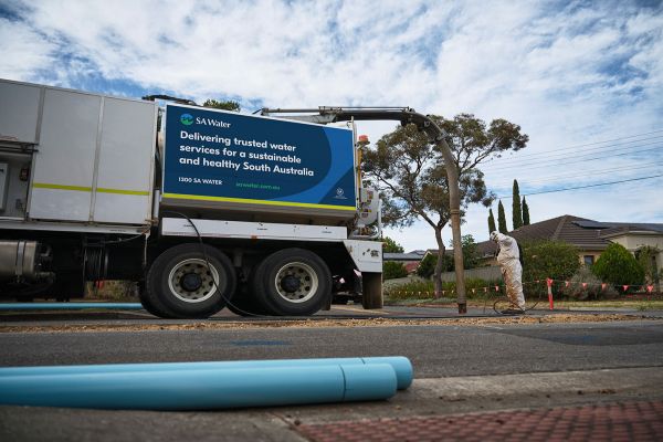 A man in white full body protective suits stands in a residential driveway. In the foreground of the photo are two pieces of blue PVC pipe, in front of an SA Water branded utility vehicle.